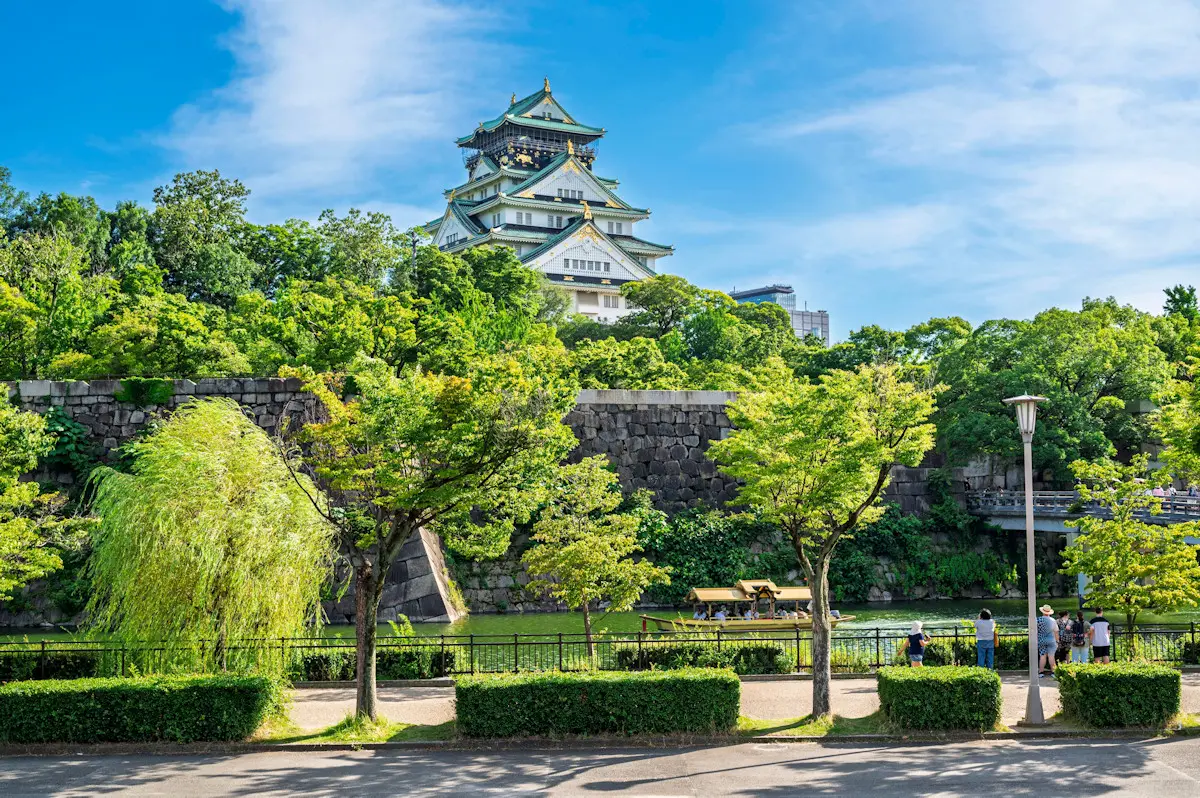 Osaka Castle is seen amongst lush green trees, set against a blue sky.