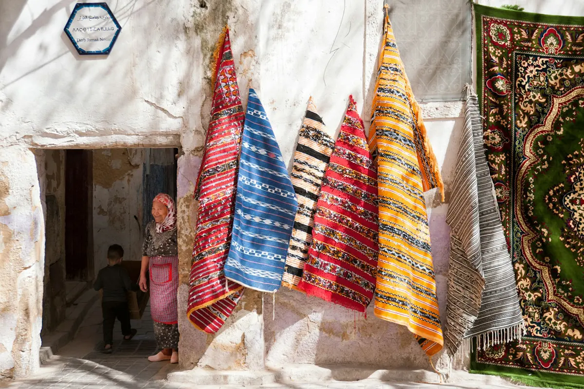 Image shows colourful rugs hanging from a wall in Fes Medina, Morocco