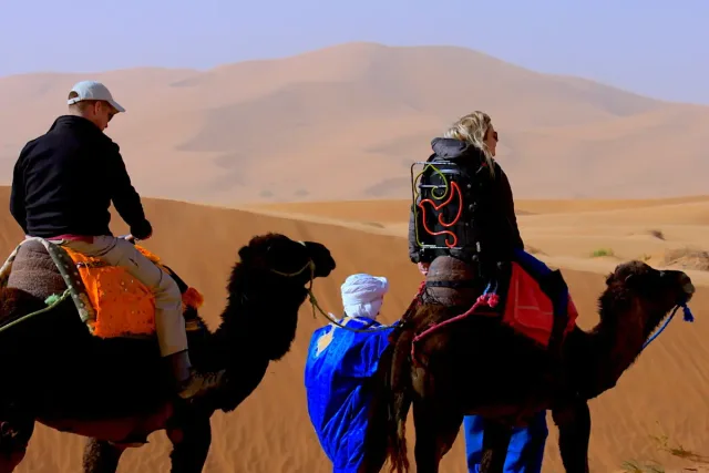 Image of two people riding camels. The woman is using an adapted saddle.
