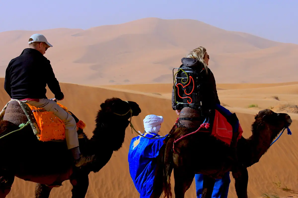 Image of two people riding camels. The woman is using an adapted saddle.