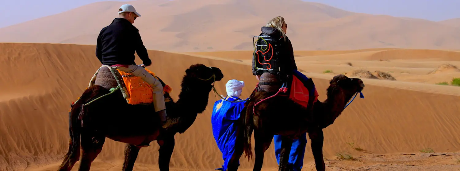 Image shows two people riding camels in Morooco. The first person is using an adapted saddle.