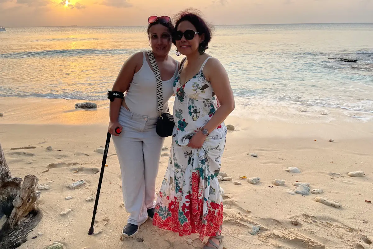 Image shows two women standing on a sandy beach. One of the women uses a walking stick